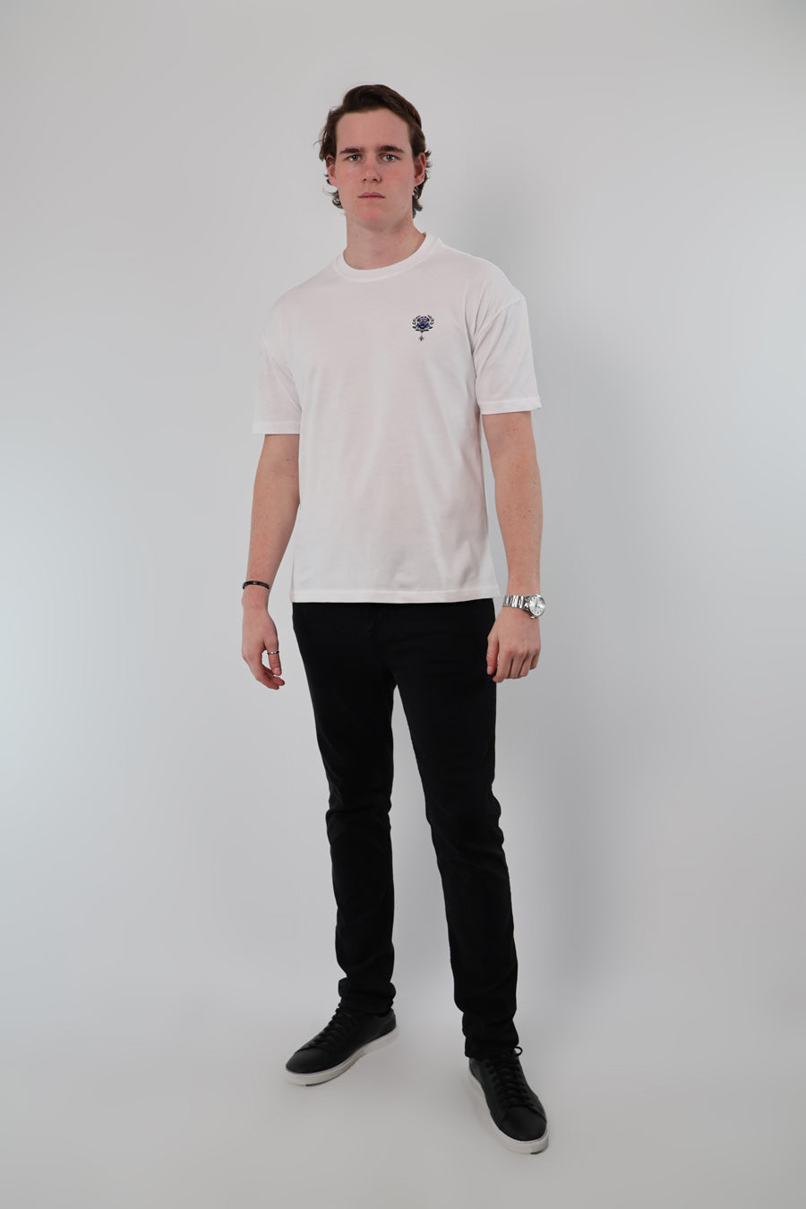 A person stands against a plain background, wearing an Aegean Rose T-shirt, black pants, and black shoes with their arms relaxed by their sides.