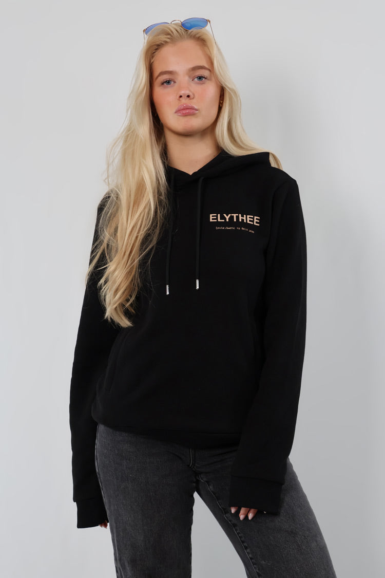 A person with long blonde hair wears the Legacy Hoodie - Unisex and gray jeans, standing against a plain light background, looking at the camera.