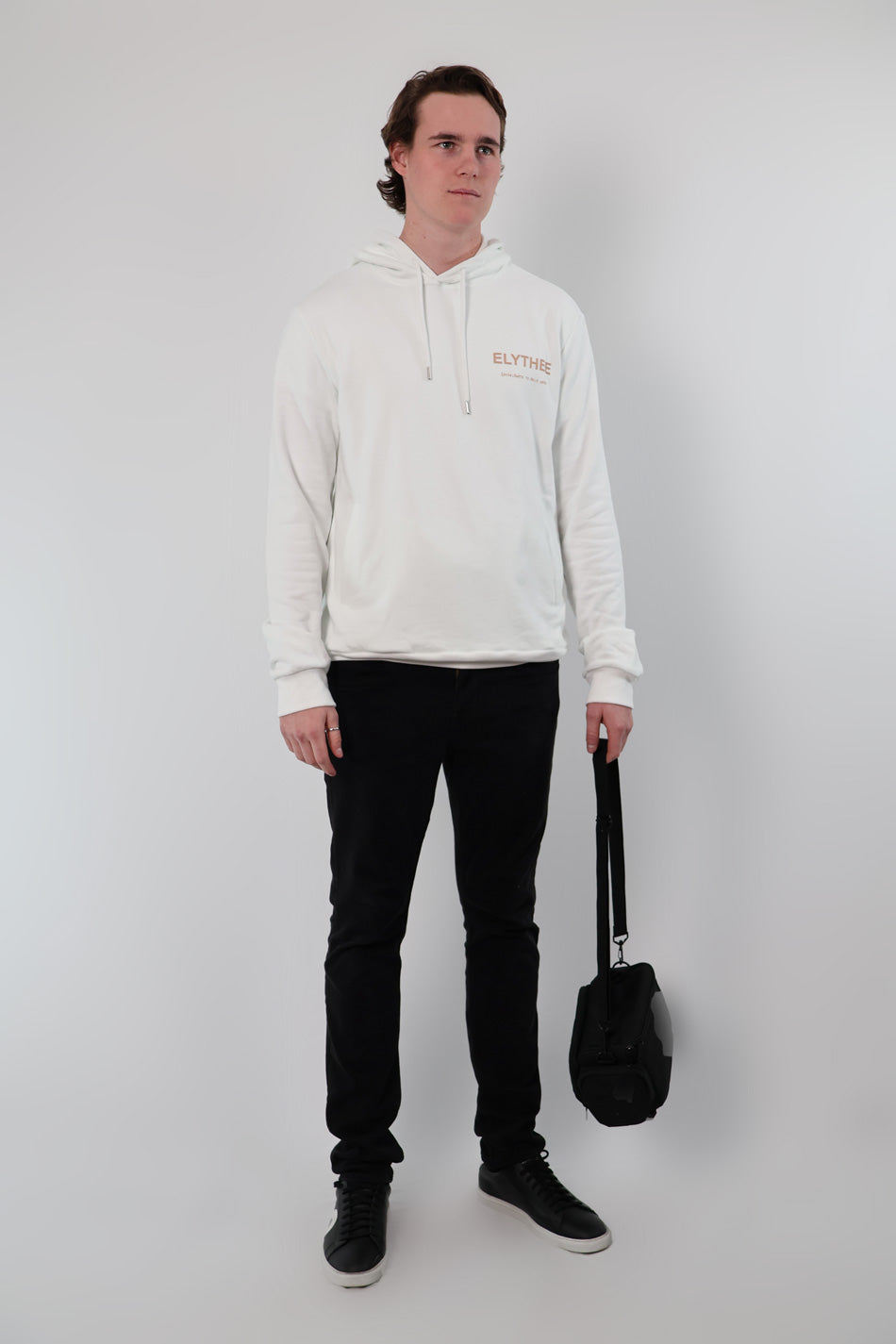 A person stands against a plain background wearing the Eternal Laurel Hoodie - Unisex in white and black pants, holding a black bag.