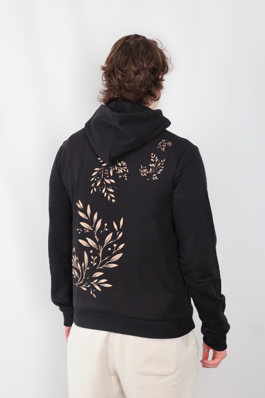 A person with long hair, seen from behind, is dressed in the Eternal Laurel Hoodie - Unisex featuring gold leafy patterns on a black background, paired with light-colored pants. Theyre standing against a plain white backdrop.
