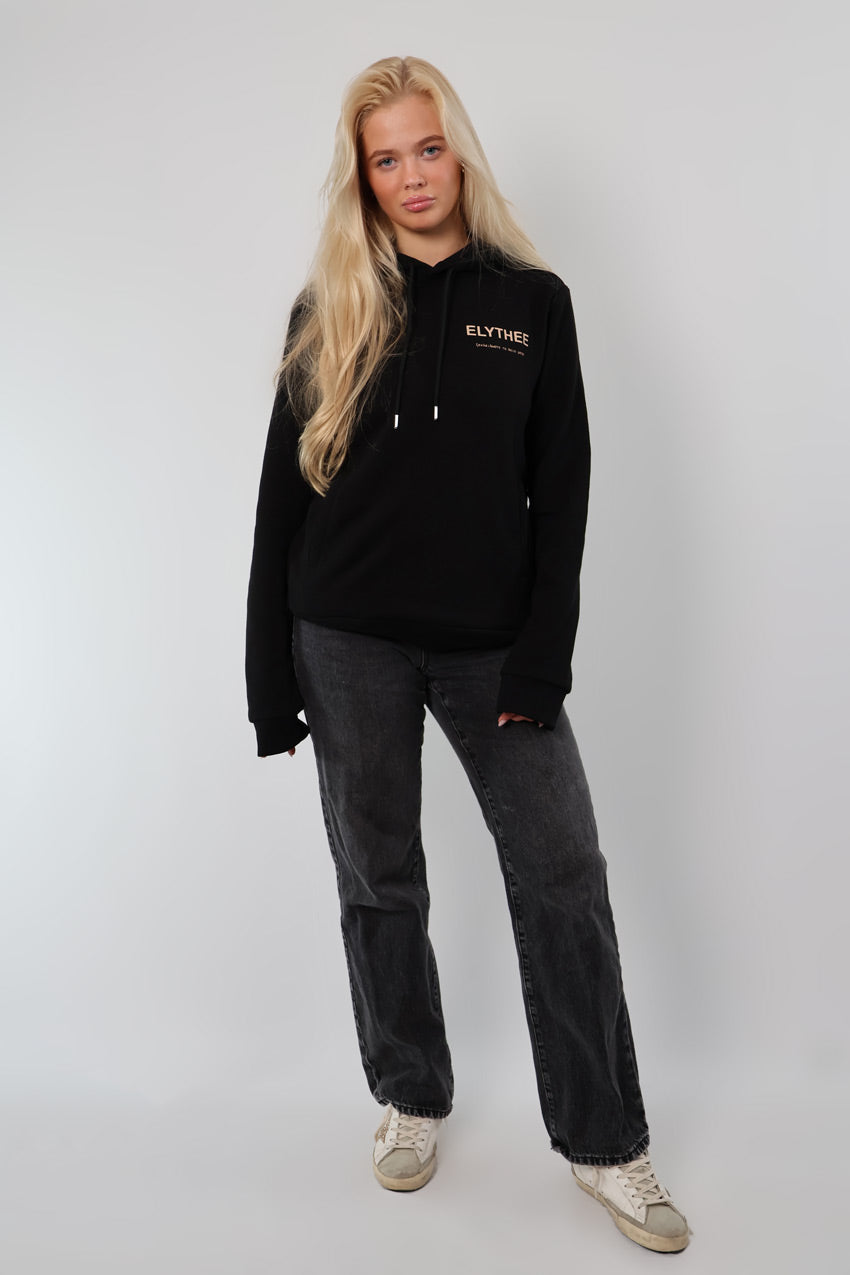 A person with long blonde hair is wearing a Legacy Hoodie - Unisex, gray jeans, and white sneakers while standing against a plain light background.