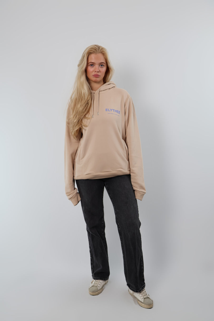 A person with long blonde hair in a Mythos Essence Hoodie - Unisex and dark jeans stands against a plain background.
