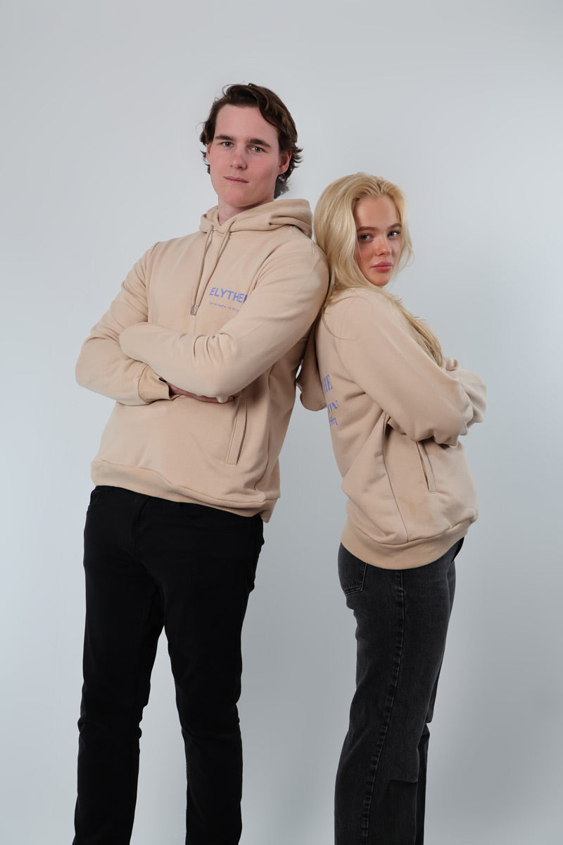 Two individuals stand back-to-back, each wearing a Mythos Essence Hoodie in beige with dark pants against a plain background.