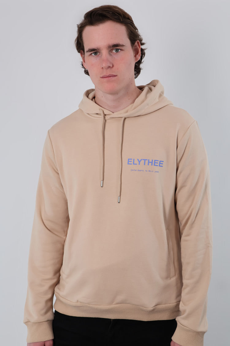 A short-haired individual wears the Mythos Essence Hoodie - Unisex, featuring ELYTHEE in blue on a beige background, posed against a plain white backdrop.