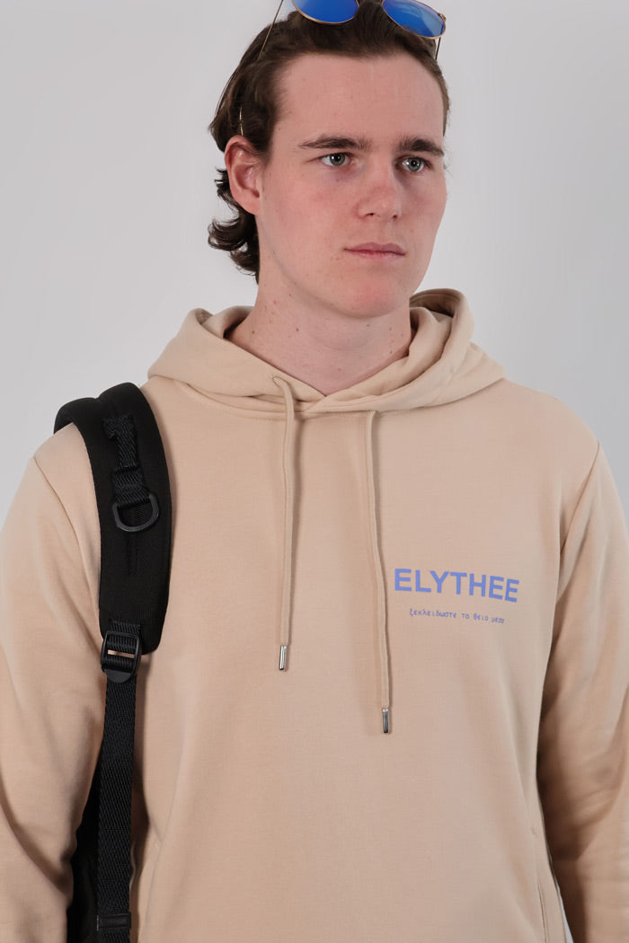 A person wearing a Mythos Essence Hoodie in beige, featuring the text ELYTHEE, and carrying a black backpack, stands against a plain background.