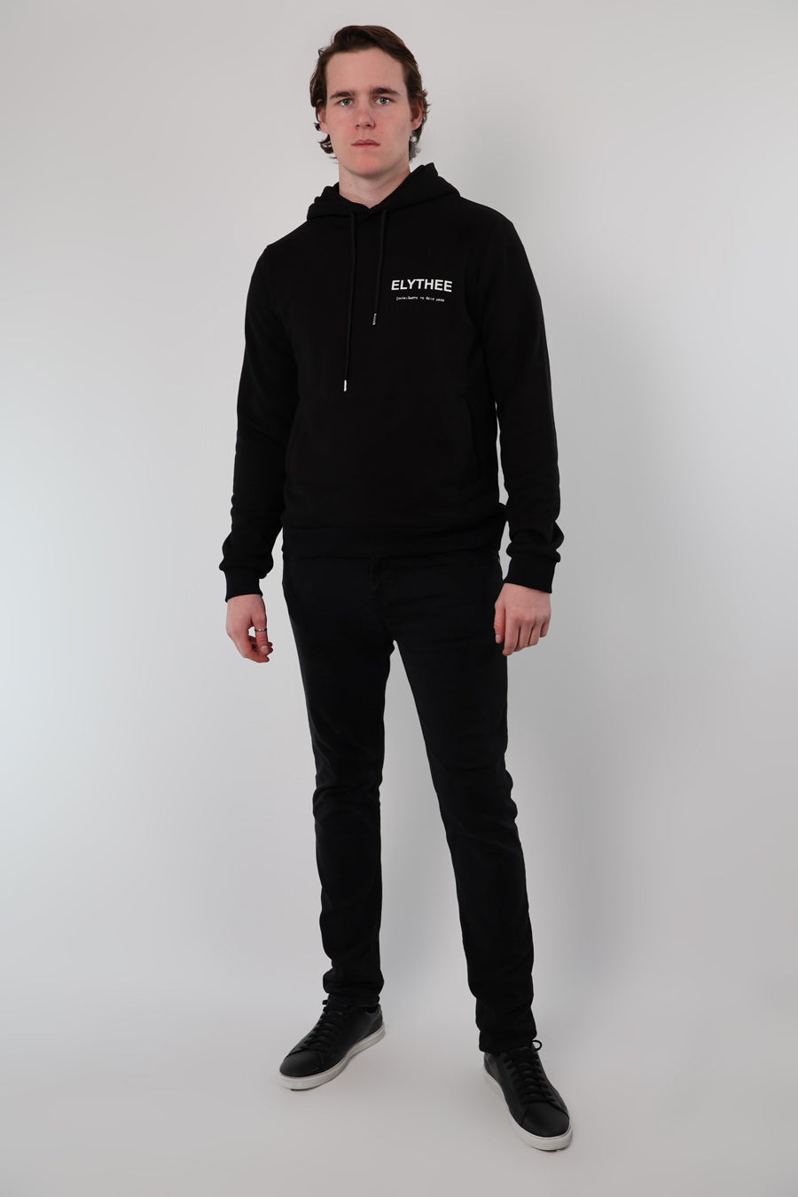 A person stands against a plain background wearing the Mythos Essence Hoodie, black pants, and black sneakers.
