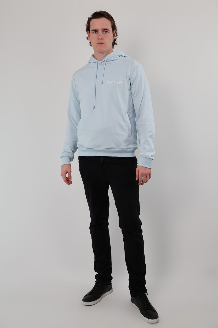 A person stands against a plain background wearing the Mythos Essence Hoodie in light blue and black pants.