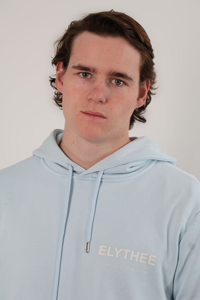 A person with wavy hair wearing a Mythos Essence Hoodie, light blue with ELYTHEE on it, stands against a plain background.