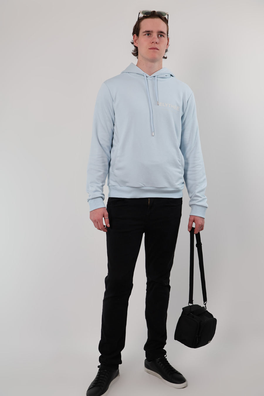 Someone wearing a Mythos Essence Hoodie in light blue, black pants, and black shoes stands holding a small black bag. Sunglasses rest on their head against a plain white background.