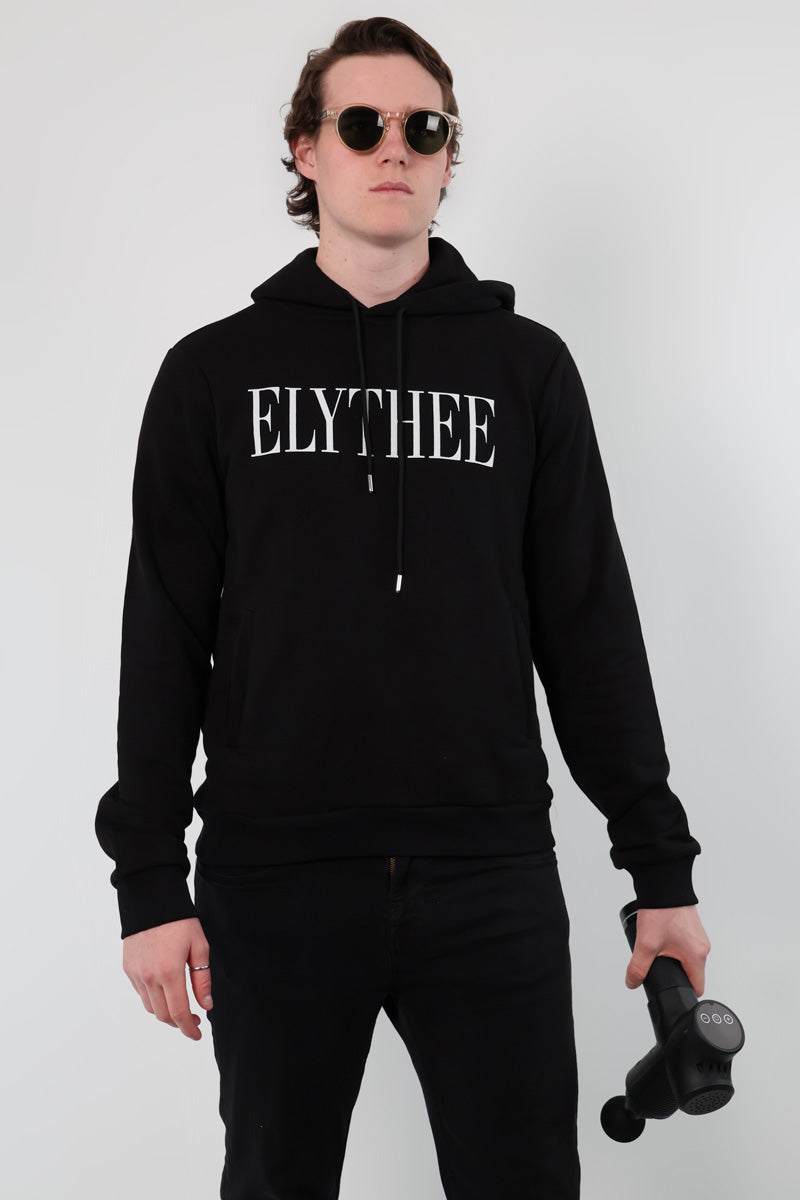 An individual in a Nectar Nomad Hoodie, wearing sunglasses and holding a massage gun, exudes confidence against a plain backdrop. The unisex hoodie enhances their enigmatic charm, capturing the essence of a true Nectar Nomad.