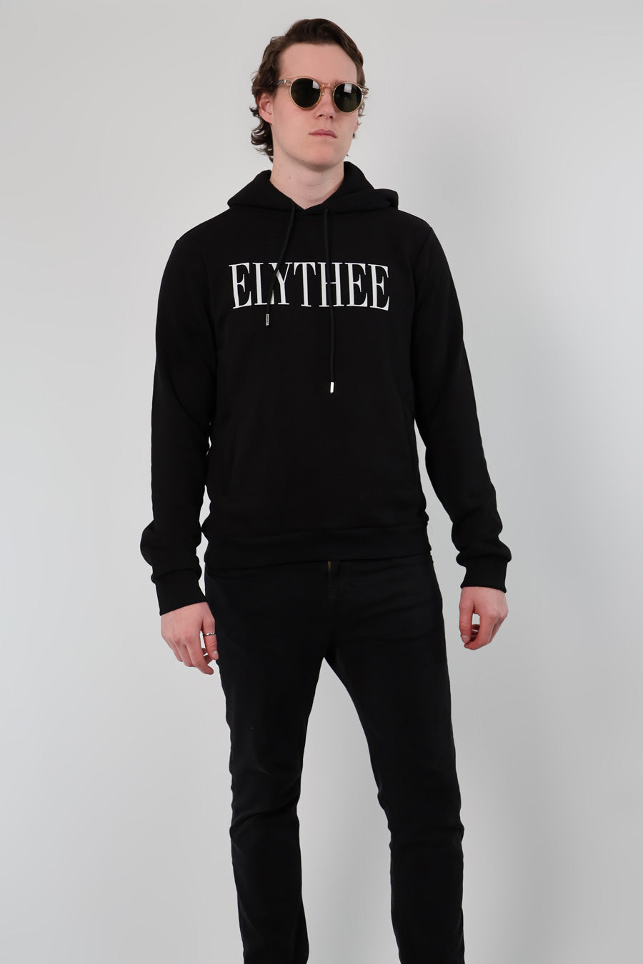 A person wearing sunglasses and a black unisex Nectar Nomad Hoodie stands against a plain light background, embodying the essence of understated style.