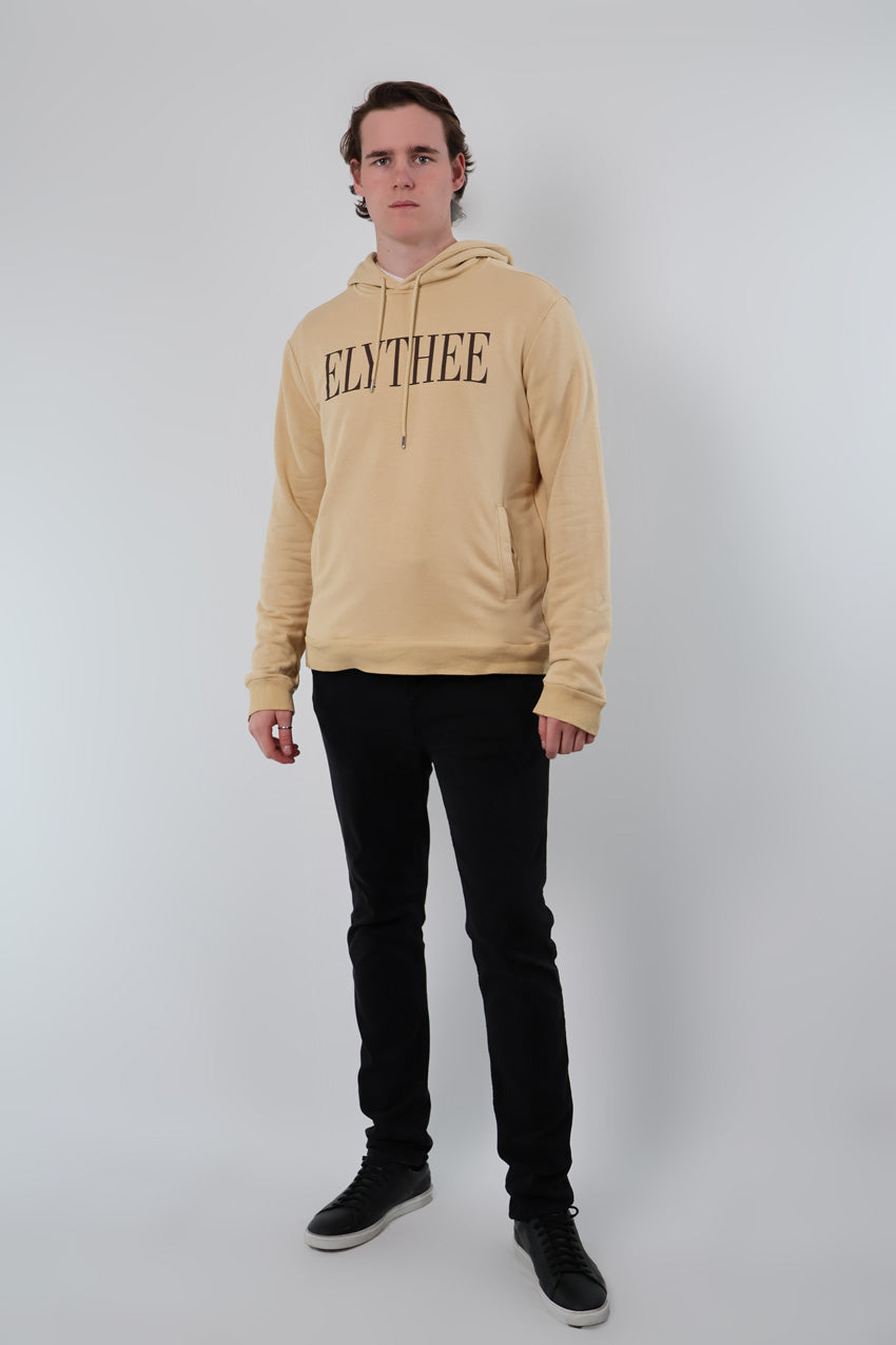A person in a beige Nectar Nomad Hoodie featuring ELYTHEE text, paired with black pants and shoes, stands against a plain backdrop, showcasing the versatile style of the Nectar Nomad.
