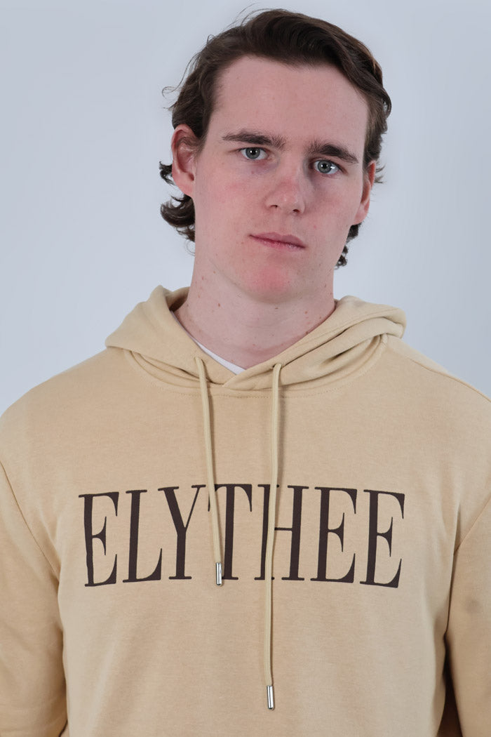 A person stands against a plain background wearing a beige Nectar Nomad Hoodie, ideal for anyone seeking comfort and style in this unisex design.