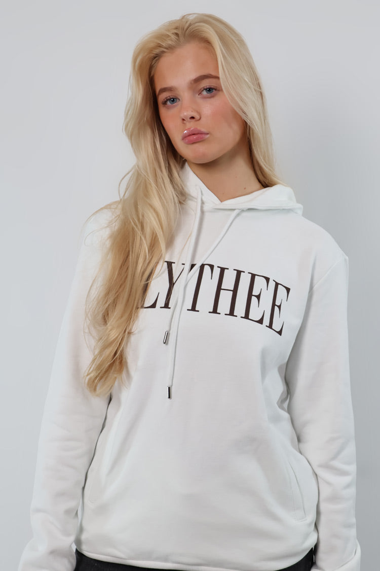 A person with long blonde hair stands gracefully against a plain backdrop, wearing a white Nectar Nomad Hoodie with LYTHEE on the front.