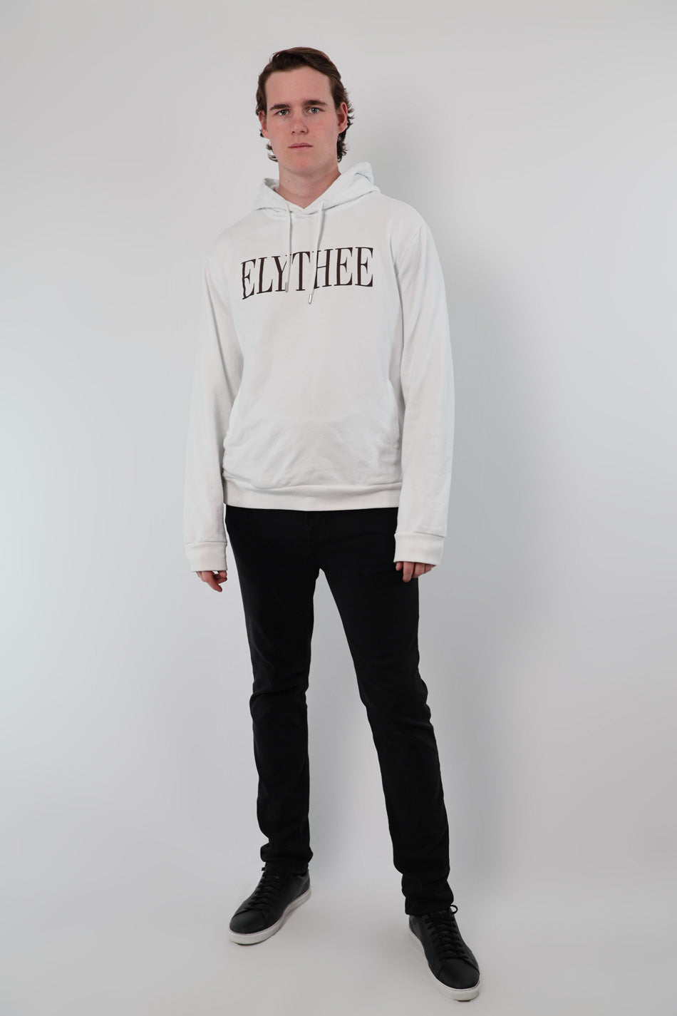 A person wears a white Nectar Nomad Hoodie with ELYTHEE prominently printed, paired with black pants, standing against a plain backdrop, effortlessly showcasing the unisex hoodie style.