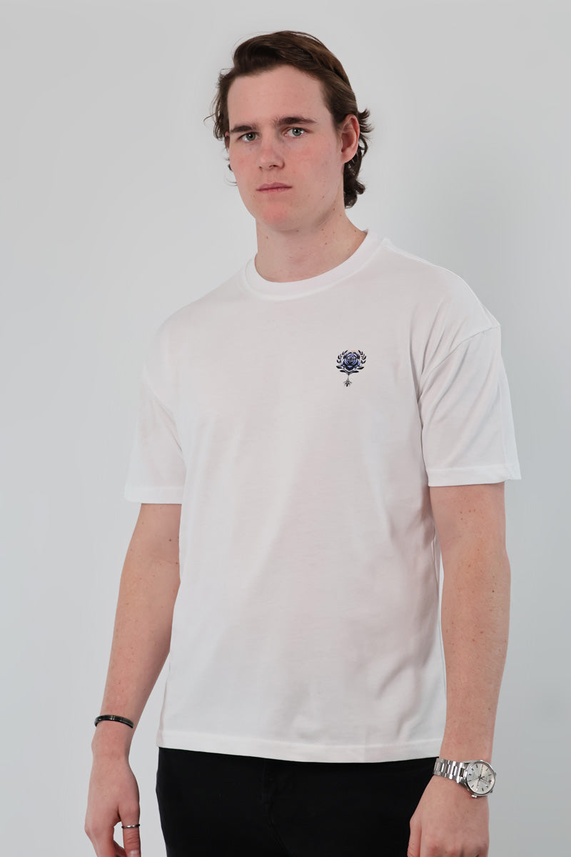 A young man is wearing the Aegean Rose T-Shirt, featuring a plain white design with a small chest logo, set against a light gray background.